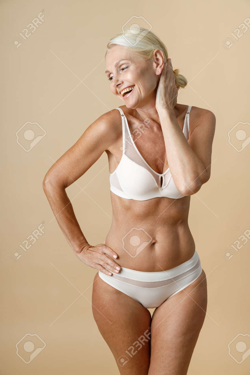 Encantadora Mujer En Interior Con Cuerpo Perfecto En Forma Sonriendo Y Tocándose La Cabeza, Parada Con Los Ojos Cerrados Aislada Sobre Un Fondo Beige Fotos, Retratos, Imágenes Y Fotografía