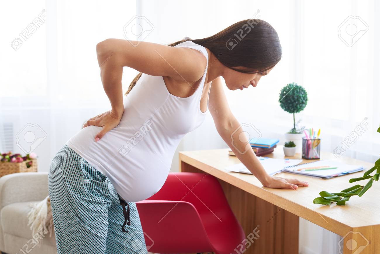 Side view of young pregnant woman suffering from backache at home, leaning on table - 86617396 Side view of young pregnant woman suffering from backache at home, leaning on table - 86617396