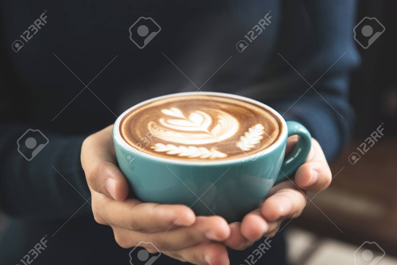 表面に美しいロゼッタラテアートパターンとコーヒーのカップを保持している女性の手 の写真素材 画像素材 Image