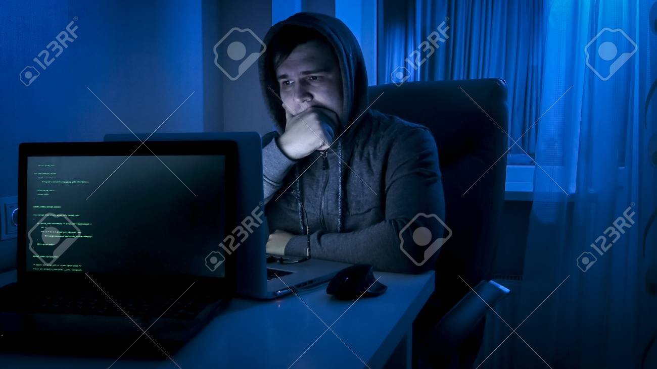 young man in hoodie looking on laptop screeen at night stock photo