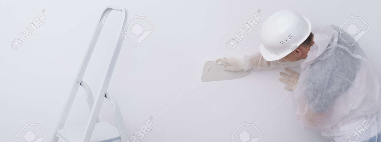 a construction worker in protective clothing and a hard hat, levels the wallpaper with a plastic spatula, to remove excess glue from the walls, there is a stepladder next to it - 169657107 a construction worker in protective clothing and a hard hat, levels the wallpaper with a plastic spatula, to remove excess glue from the walls, there is a stepladder next to it - 169657107
