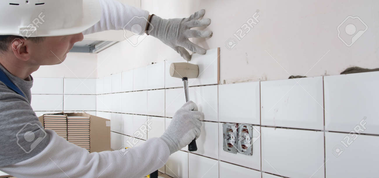 a builder in a protective helmet, using a rubber hammer, corrects the correct installation of tiles - 169657167 a builder in a protective helmet, using a rubber hammer, corrects the correct installation of tiles - 169657167