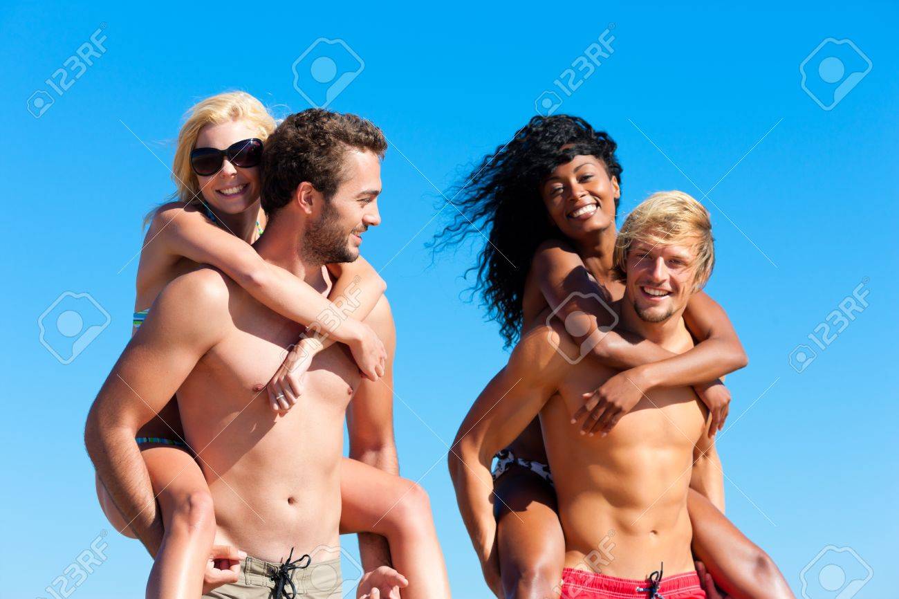 Cuatro Amigos, Hombres Y Mujeres En La Playa Con Un Montón De Diversión,  Los Hombres Que Llevaban Las Mujeres Empacar De Nuevo Fotos, retratos,  imágenes y fotografía de archivo libres de derecho.