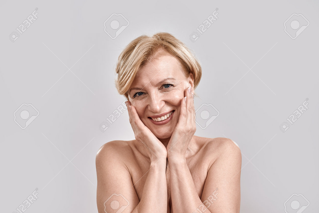 Portrait Of Beautiful Naked Middle Aged Woman Looking Excited, Smiling At  Camera, Touching Her Cheeks While Posing Isolated Against Grey Background.  Beauty, Skincare Concept Stock Photo, Picture and Royalty Free Image. Image