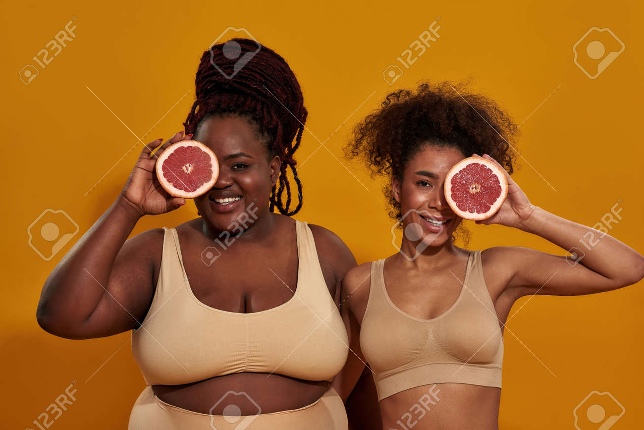 De Estudio De Alegres Mujeres Afroamericanas En Ropa Interior Sonriendo A La Cámara, Cubriendo Un Ojo Con La Mitad Toronja, Posando Aisladas Sobre Fondo Naranja Fotos, Retratos, Imágenes
