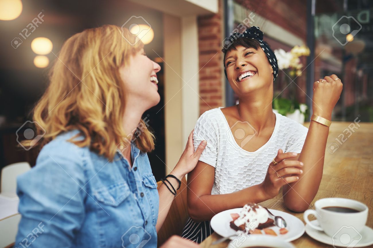 Two multiethnic young female friends enjoying coffee together in a restaurant laughing and joking while touching to display affection - 46626116 Two multiethnic young female friends enjoying coffee together in a restaurant laughing and joking while touching to display affection - 46626116