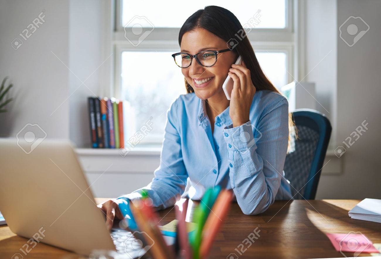 Single happy female business owner with smile and eyeglasses on phone and working on laptop computer at desk with bright window in background - 54383974 Single happy female business owner with smile and eyeglasses on phone and working on laptop computer at desk with bright window in background - 54383974