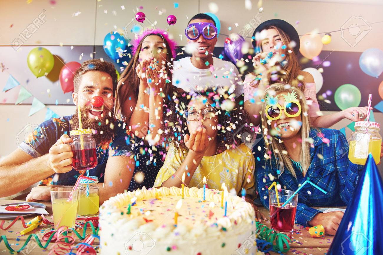 Group of six male and female festive partygoers in front of large white frosting covered cake surrounded by confetti in room - 64785334 Group of six male and female festive partygoers in front of large white frosting covered cake surrounded by confetti in room - 64785334