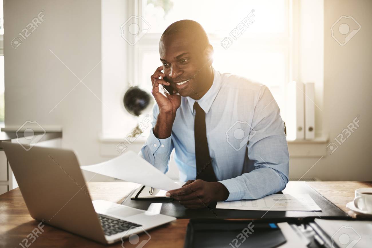 オフィスの机に座りながら 携帯電話でクライアントと話している若いアフリカのビジネスマンの笑顔 の写真素材 画像素材 Image