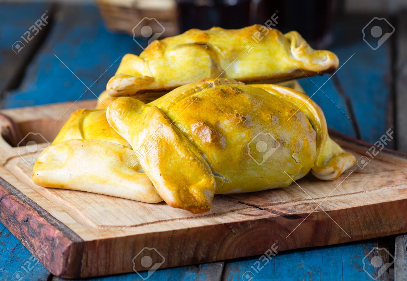 Empanadas. Latin American chilean food. Typical traditional Chilean empanadas with meat, onion, eggs and olives on mapuche rustic cutting board on wooden blue background - 65537918 Empanadas. Latin American chilean food. Typical traditional Chilean empanadas with meat, onion, eggs and olives on mapuche rustic cutting board on wooden blue background - 65537918
