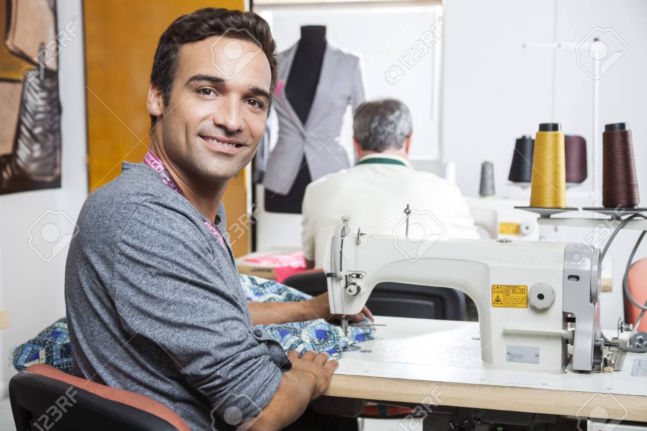 バック グラウンドで同僚と工場でミシンを使用して笑みを浮かべて男性仕立て屋の側ビュー肖像画 の写真素材 画像素材 Image