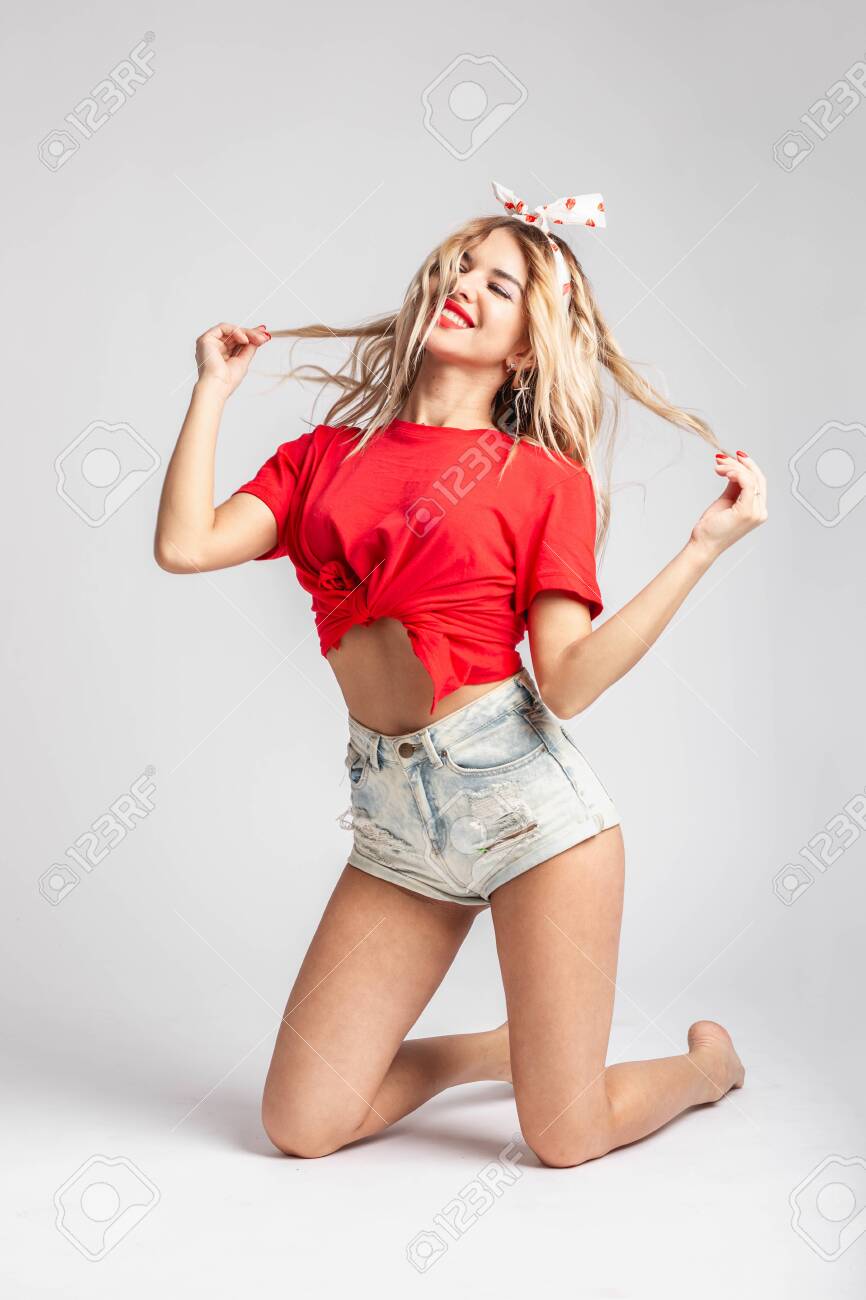 Young blond girl with a ribbon on her head dressed in short denim shorts and a red t-shirt poses sitting on the floor against a wall in the studio - 123174368 Young blond girl with a ribbon on her head dressed in short denim shorts and a red t-shirt poses sitting on the floor against a wall in the studio - 123174368