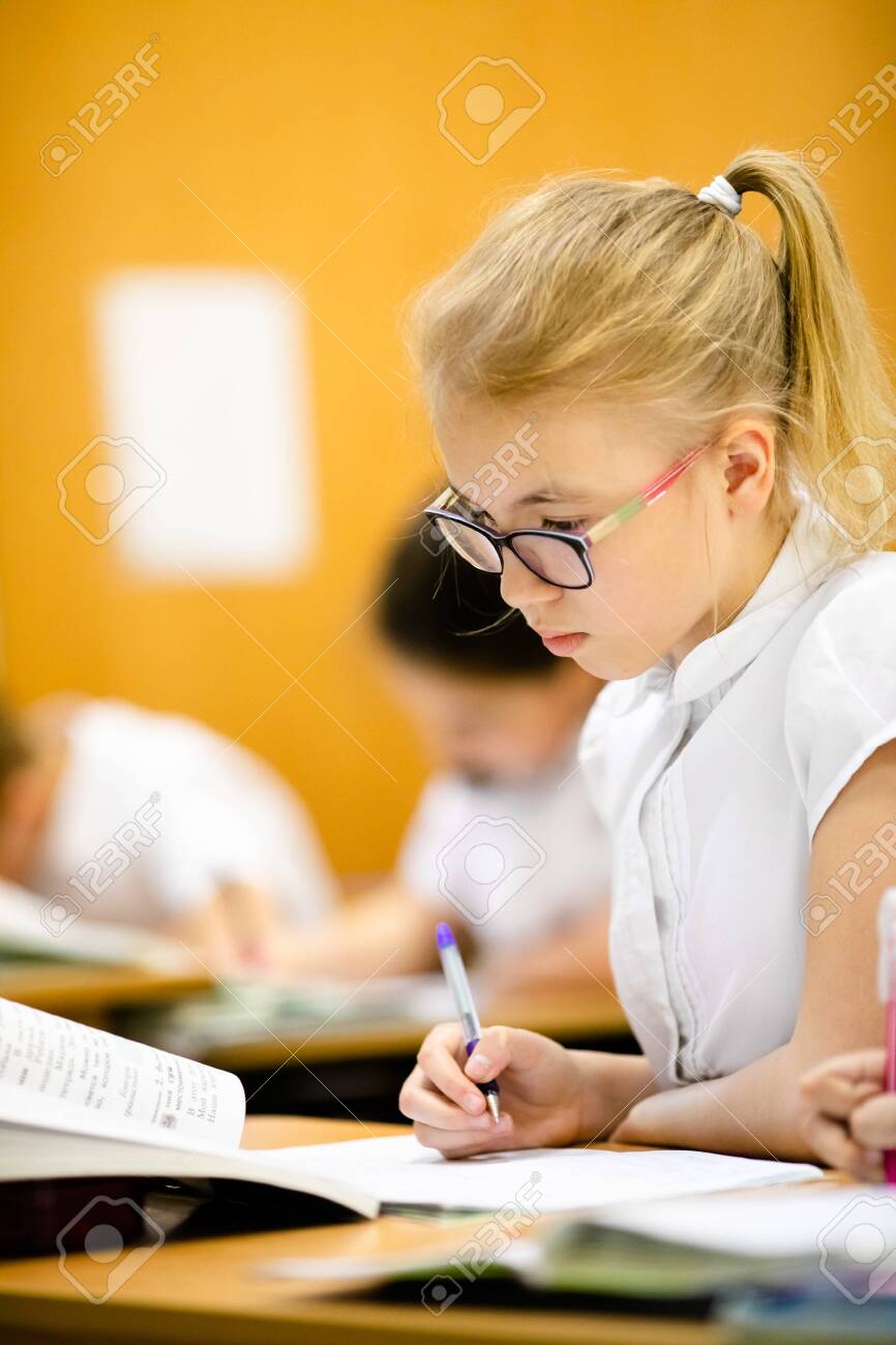 Smiling scholar girl sitting with other children in classroom and writing on textbook. Happy student doing homework at elementary school. Young schoolgirl feeling confident while writing on notebook. - 141784059 Smiling scholar girl sitting with other children in classroom and writing on textbook. Happy student doing homework at elementary school. Young schoolgirl feeling confident while writing on notebook. - 141784059