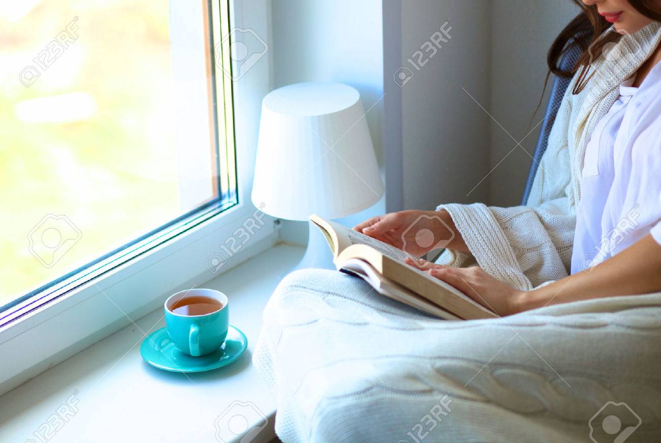 彼女のリビング ルームの読書でのんびりとコーヒーやお茶を飲んだの窓の近くに若い女性自宅が座っています の写真素材 画像素材 Image