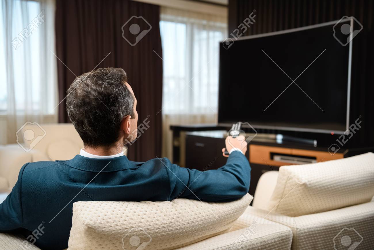 Businessman in formal suit sitting on sofa in hotel room and watching television - 102318792 Businessman in formal suit sitting on sofa in hotel room and watching television - 102318792