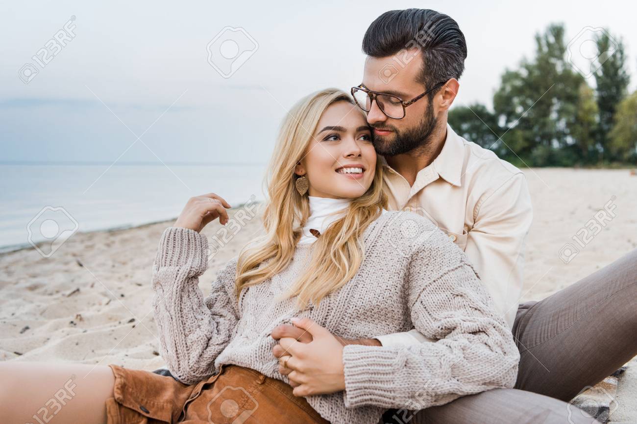Smiling girlfriend and boyfriend in autumn outfit sitting and hugging on beach - 110357948 Smiling girlfriend and boyfriend in autumn outfit sitting and hugging on beach - 110357948