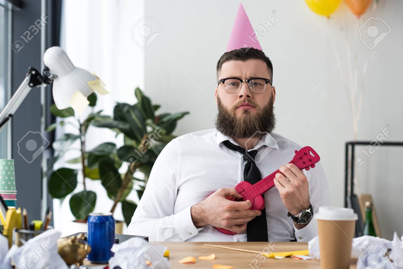 portrait of businessman with party cone on head and toy guitar at workplace in office - 112756483 portrait of businessman with party cone on head and toy guitar at workplace in office - 112756483