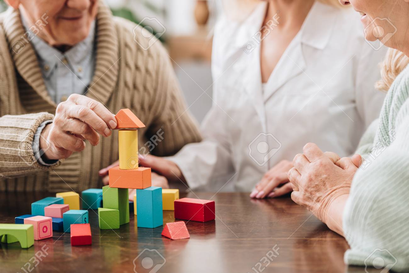 cropped view of caregiver sitting with retired man and woman and playing with wooden toys - 117439376 cropped view of caregiver sitting with retired man and woman and playing with wooden toys - 117439376