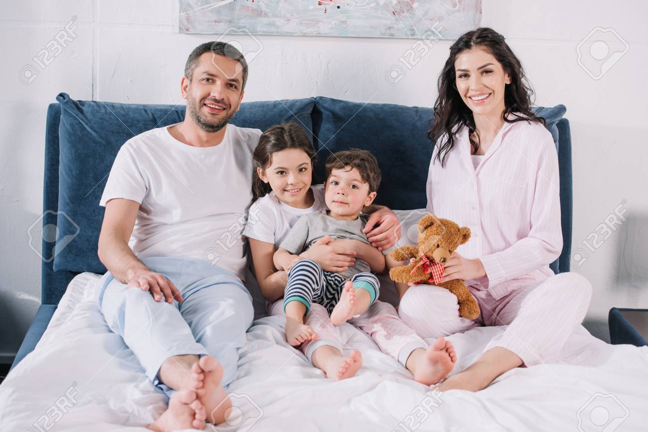 cheerful family with barefoot sitting on bed and smiling while looking at camera - 122853722 cheerful family with barefoot sitting on bed and smiling while looking at camera - 122853722