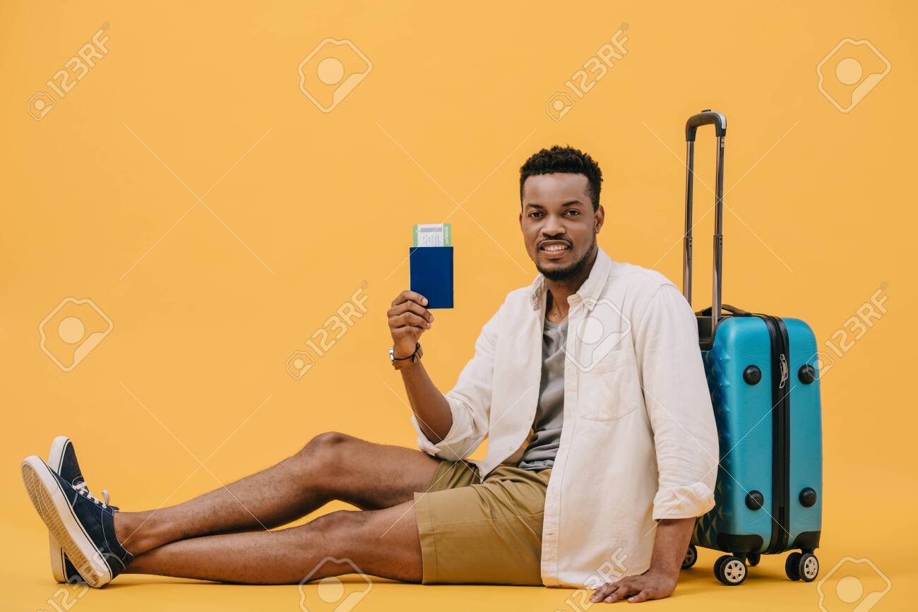 130420668-happy-african-american-man-holding-passport-with-air-ticket-and-sitting-near-luggage-on-orange.jpg