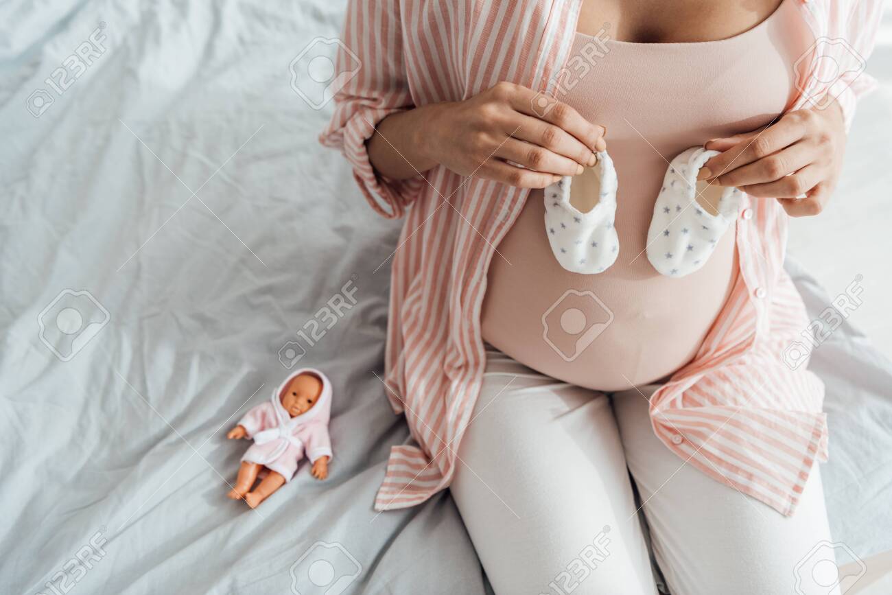 cropped view of pregnant woman sitting on bed and holding baby shoes - 131262653 cropped view of pregnant woman sitting on bed and holding baby shoes - 131262653
