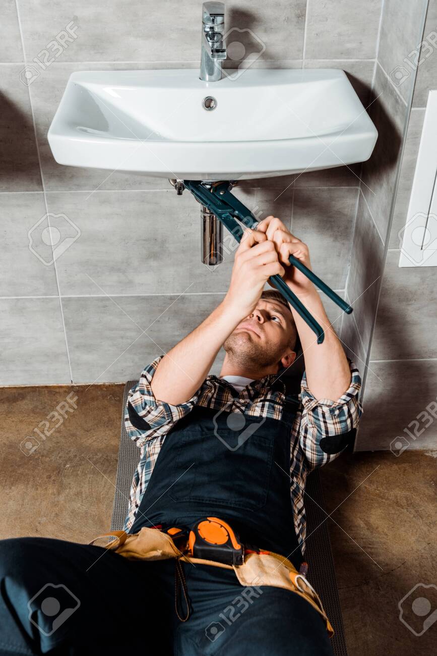 Focused Installer Holding Slip Joint Pliers Near Pipe In Bathroom Stock Photo Focused Installer Holding Slip Joint Pliers Near Pipe In Bathroom Stock Photo