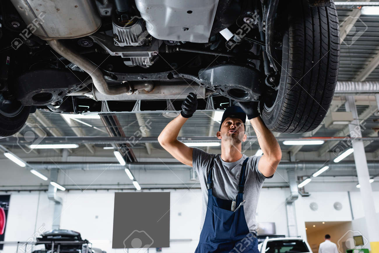 low angle view of technician in cap and overalls repairing car in service station - 152273748 low angle view of technician in cap and overalls repairing car in service station - 152273748