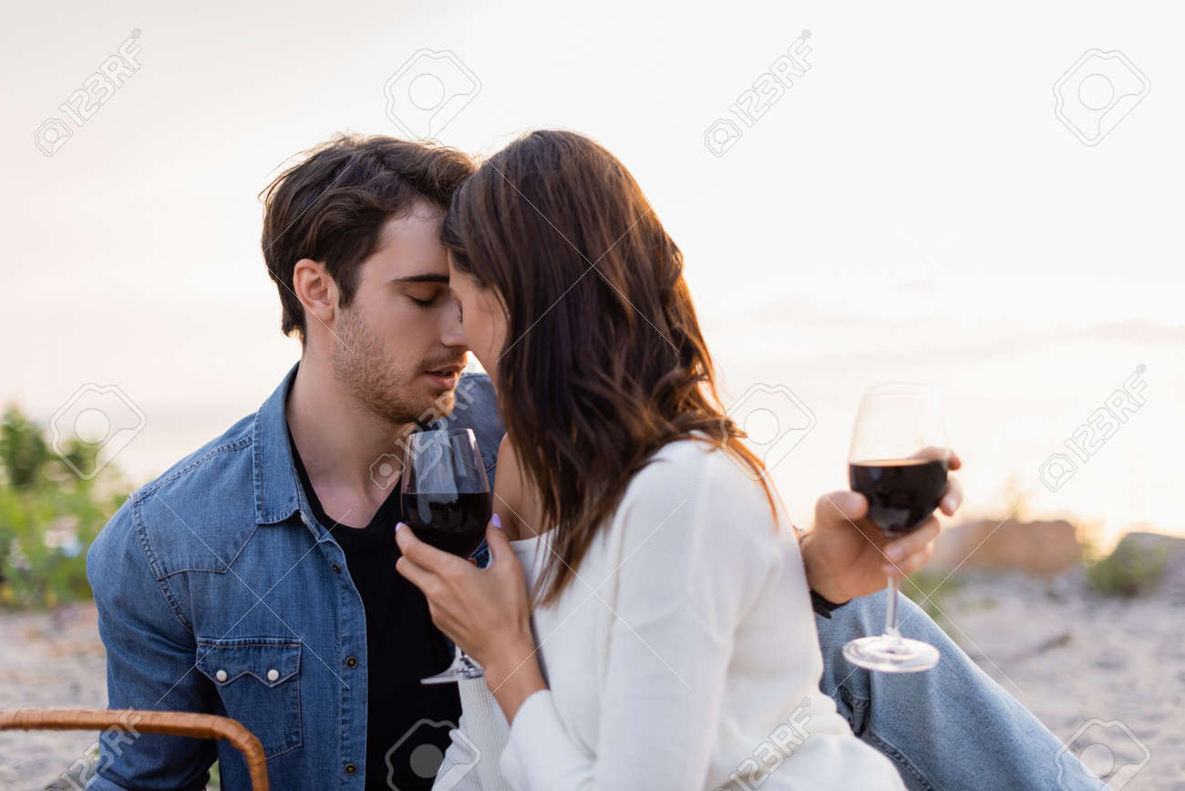 Young couple with glasses of wine kissing on beach at sunset - 156592425 Young couple with glasses of wine kissing on beach at sunset - 156592425