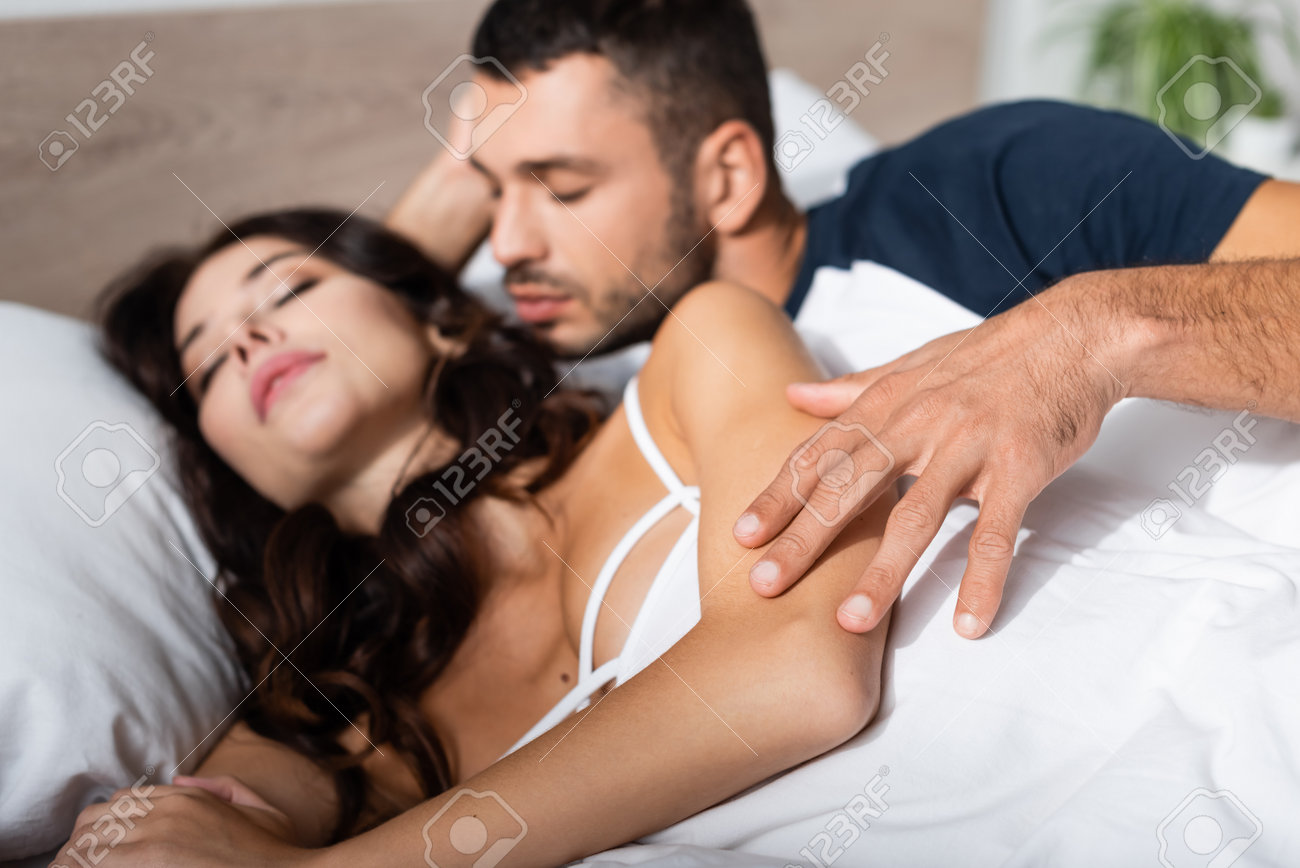 Tender moment of man gently touching woman's arm as she lies relaxed in bed, eyes closed in dreamy bliss