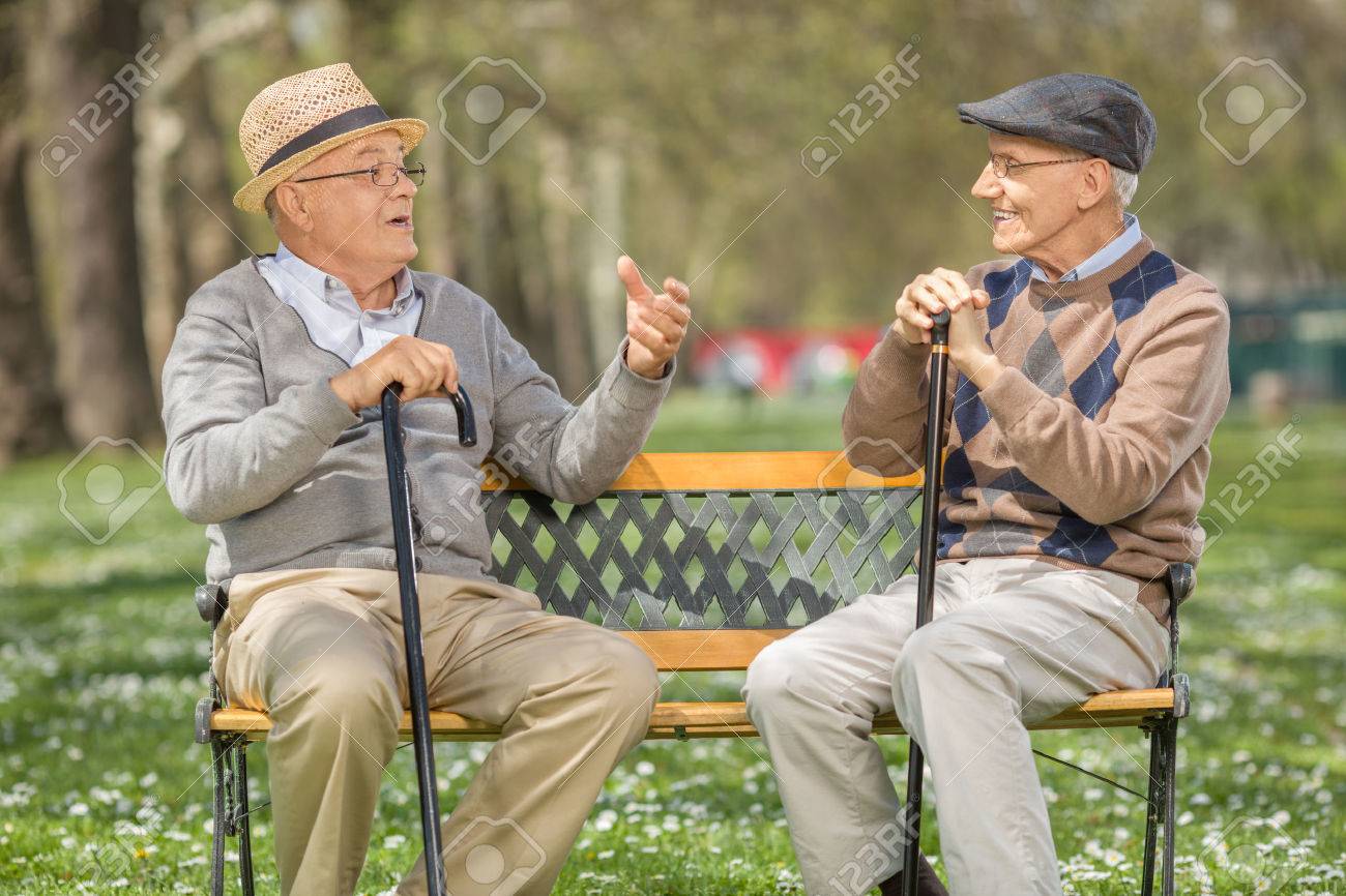 Two cheerful senior gentlemen talking to each other seated on a bench in a park - 55832108 Two cheerful senior gentlemen talking to each other seated on a bench in a park - 55832108