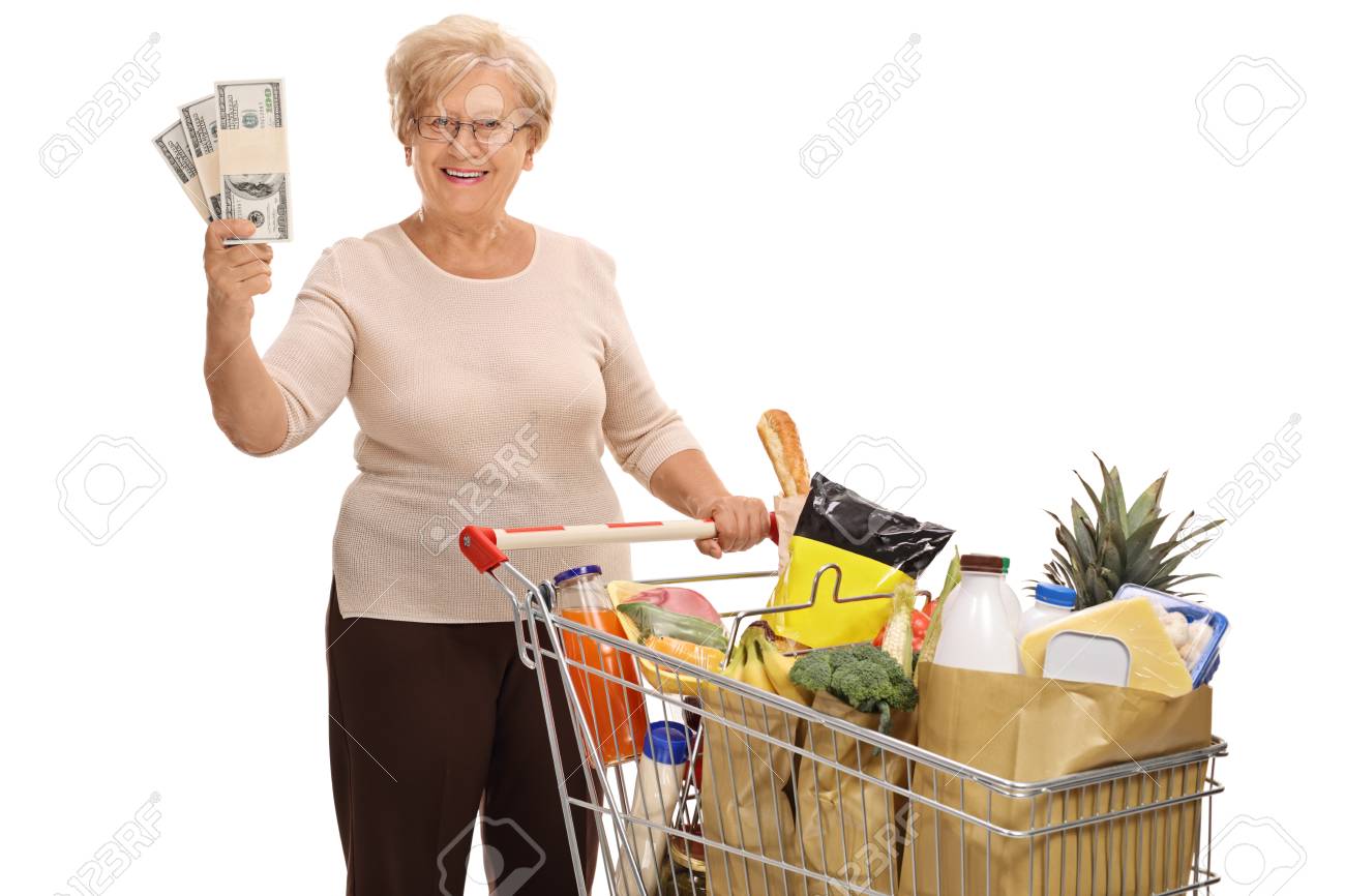 食料品 白い背景で隔離のお金のスタックのショッピング カートでポーズをとって幸せな高齢女 の写真素材 画像素材 Image