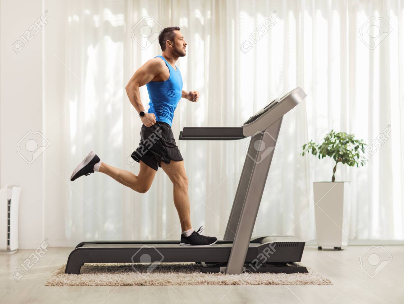 Full length profile shot of a young man running on a treadmill at home - 129384973 Full length profile shot of a young man running on a treadmill at home - 129384973