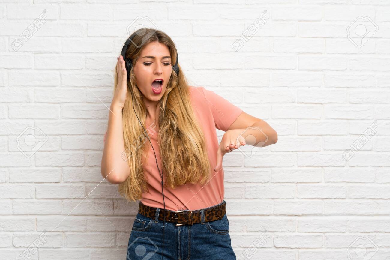 Young blonde woman over white brick wall listening to music with headphones - 127037500 Young blonde woman over white brick wall listening to music with headphones - 127037500