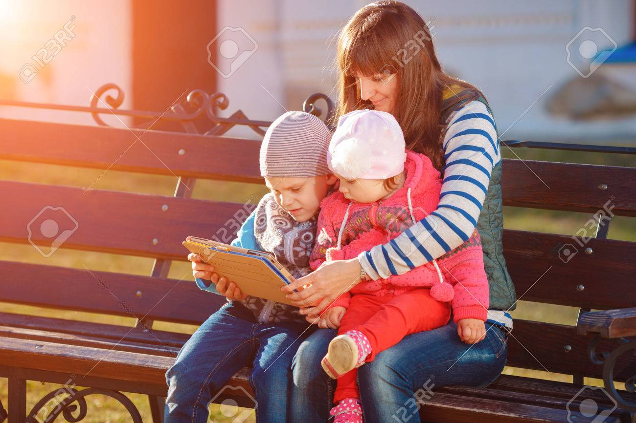 Happy family of three, mother, son and daughter sitting on a bench in the afternoon in the city park and are holding a tablet, a beautiful spring weather, the sun is shining - 39283566 Happy family of three, mother, son and daughter sitting on a bench in the afternoon in the city park and are holding a tablet, a beautiful spring weather, the sun is shining - 39283566