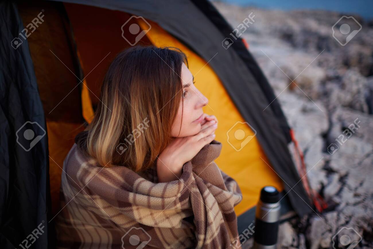 Handsome woman sitting with hand under chin, on stone beach, looking at sunset. - 122905199 Handsome woman sitting with hand under chin, on stone beach, looking at sunset. - 122905199