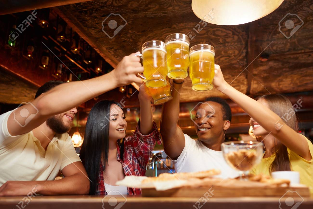 Four boys and two girls having fun together, raising beer above table. - 107927664 Four boys and two girls having fun together, raising beer above table. - 107927664