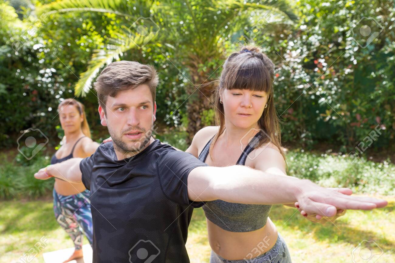 Focused yoga instructor helping newby to cope with warrior pose. Man doing yoga outdoors, woman adjusting his hands. Training yoga concept - 123848391 Focused yoga instructor helping newby to cope with warrior pose. Man doing yoga outdoors, woman adjusting his hands. Training yoga concept - 123848391