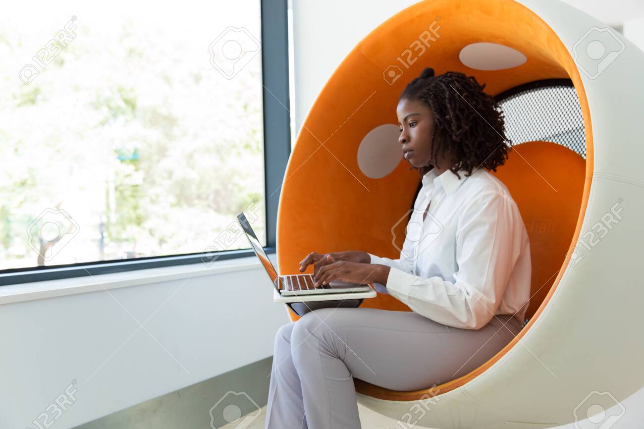 Serious young female professional working on computer in office rest room. Black business woman sitting in interactive chair, using laptop, typing and looking at screen. Busy professional concept - 130048374 Serious young female professional working on computer in office rest room. Black business woman sitting in interactive chair, using laptop, typing and looking at screen. Busy professional concept - 130048374