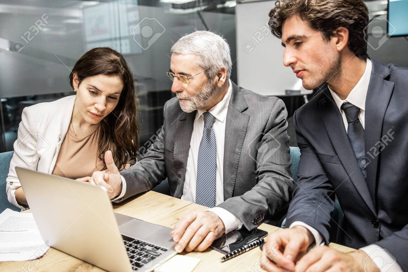 Confident business team looking at laptop. Group of office employees sitting at table and discussing new project. Business, teamwork concept - 145407937 Confident business team looking at laptop. Group of office employees sitting at table and discussing new project. Business, teamwork concept - 145407937