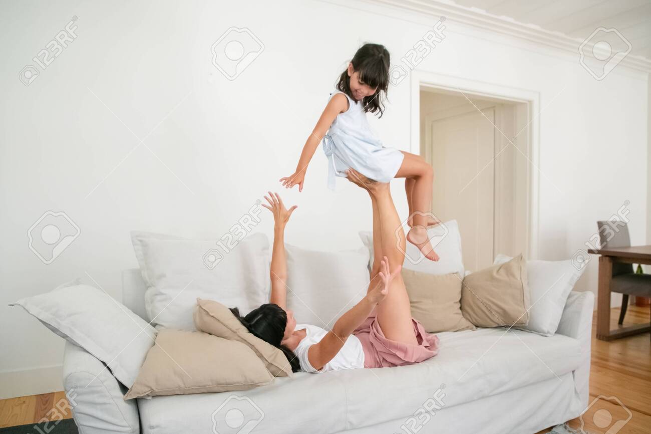 Cheerful Mother Lying On Sofa And Holding Daughter With Legs. Young  Caucasian Mom Playing Game With Adorable Little Girl And Spreading Hands In  Living Room. Motherhood, Family Time And Leisure Concept Stock
