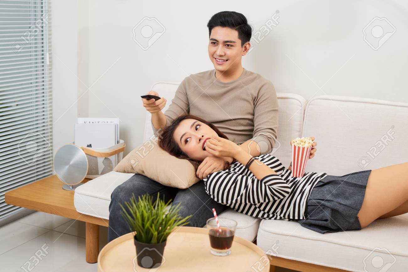 Young couple in love sitting on a brown sofa and watching television in the living room. - 107467422 Young couple in love sitting on a brown sofa and watching television in the living room. - 107467422