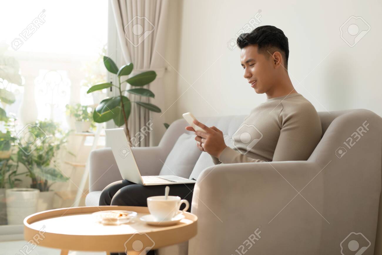 Young Asian man relaxing at home on a sofa with his laptop computer chatting on his mobile while reading the screen of the laptop with a smile - 140436752 Young Asian man relaxing at home on a sofa with his laptop computer chatting on his mobile while reading the screen of the laptop with a smile - 140436752