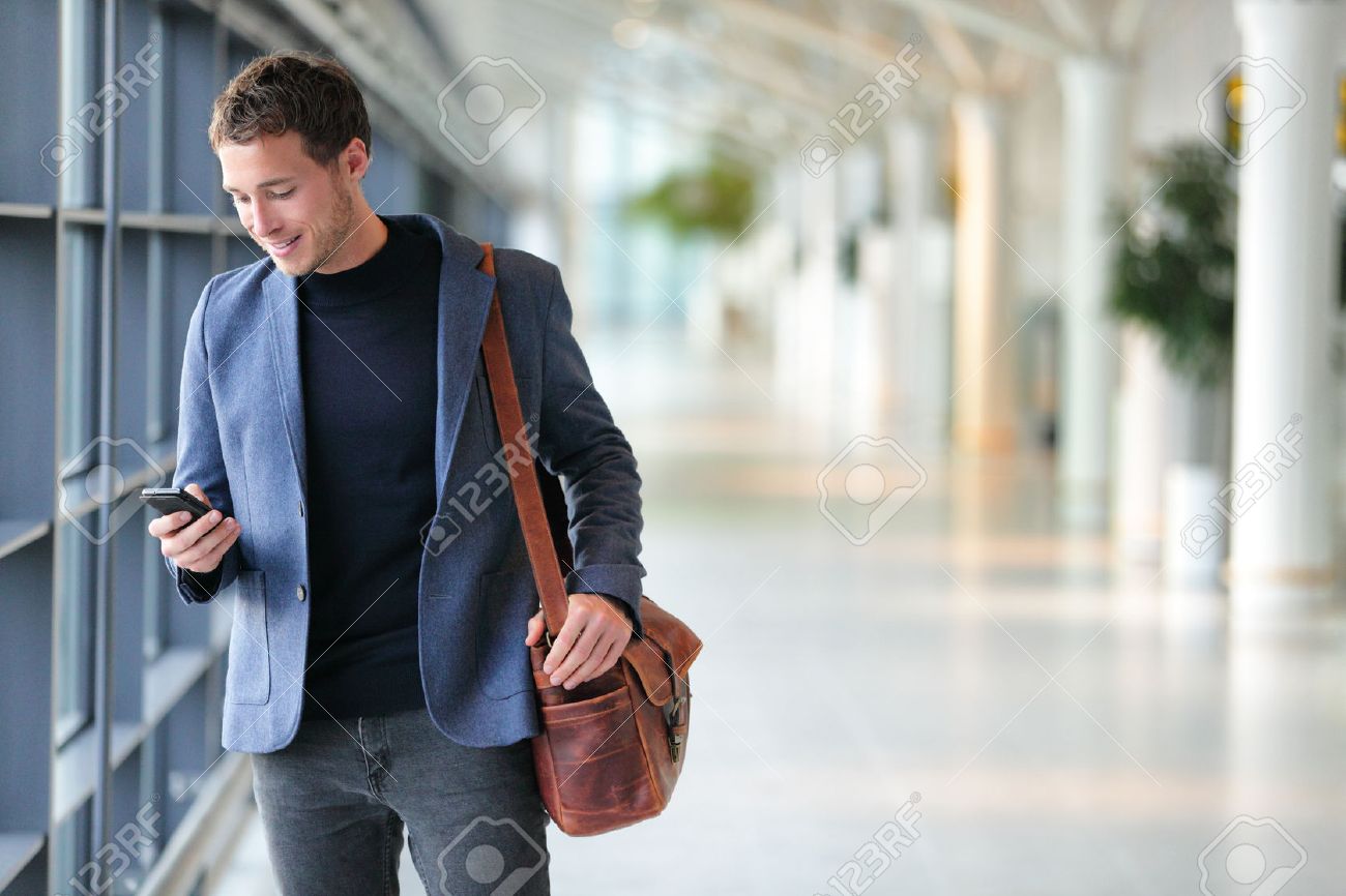 Business man using mobile phone app in airport. Young business professional man texting smartphone walking inside office building or airport terminal. Handsome man wearing stylish suit jacket indoors. - 65498953 Business man using mobile phone app in airport. Young business professional man texting smartphone walking inside office building or airport terminal. Handsome man wearing stylish suit jacket indoors. - 65498953