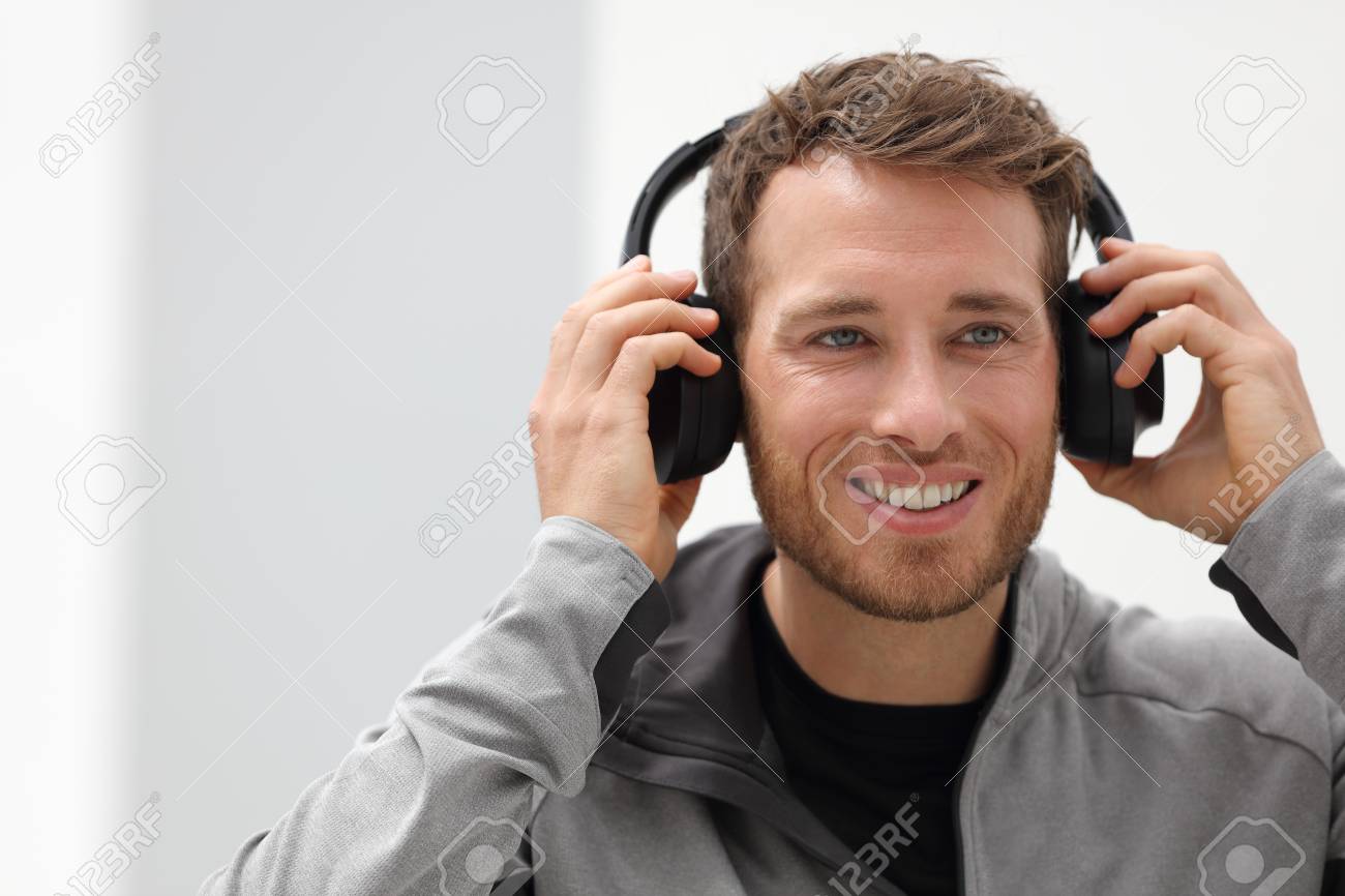 Man putting on headphones to listen to music mobile phone app. Happy smiling young urban person wearing headset sing smartphone mobile app listening to songs in living room at home. - 90920641 Man putting on headphones to listen to music mobile phone app. Happy smiling young urban person wearing headset sing smartphone mobile app listening to songs in living room at home. - 90920641