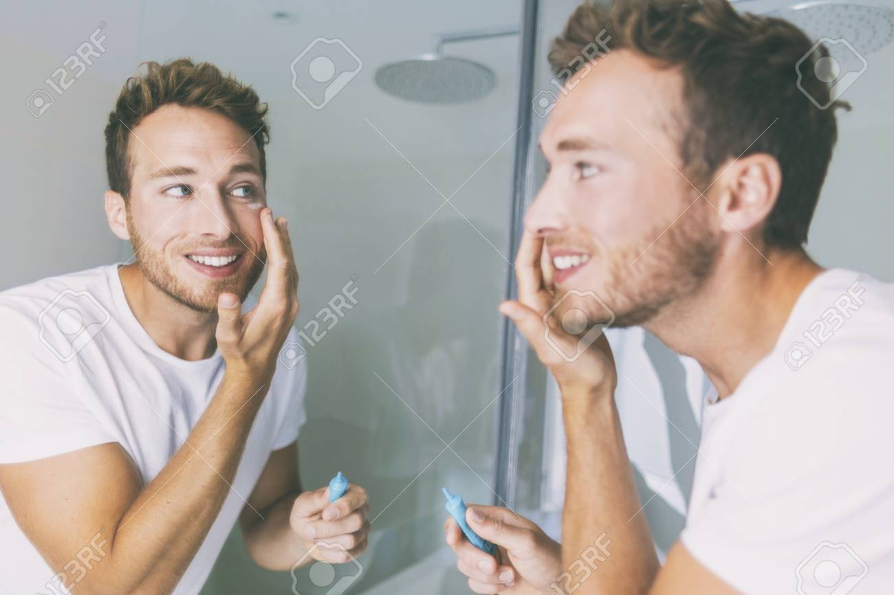 Man putting skincare facial treatment cream on face. Anti-aging skin care product. Male beauty morning routine at home lifestyle. Guy looking in bathroom mirror applying moisturizer under eyes. - 115599108 Man putting skincare facial treatment cream on face. Anti-aging skin care product. Male beauty morning routine at home lifestyle. Guy looking in bathroom mirror applying moisturizer under eyes. - 115599108