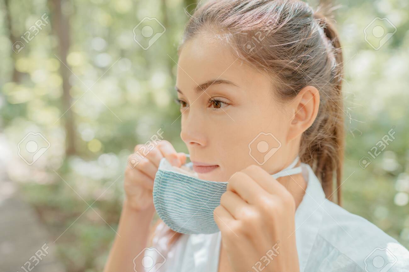 Asian beauty woman putting on mask on face. Wearing corona virus prevention masks for long hours are creating irritation, skin problems, acne pimples. - 154570627 Asian beauty woman putting on mask on face. Wearing corona virus prevention masks for long hours are creating irritation, skin problems, acne pimples. - 154570627