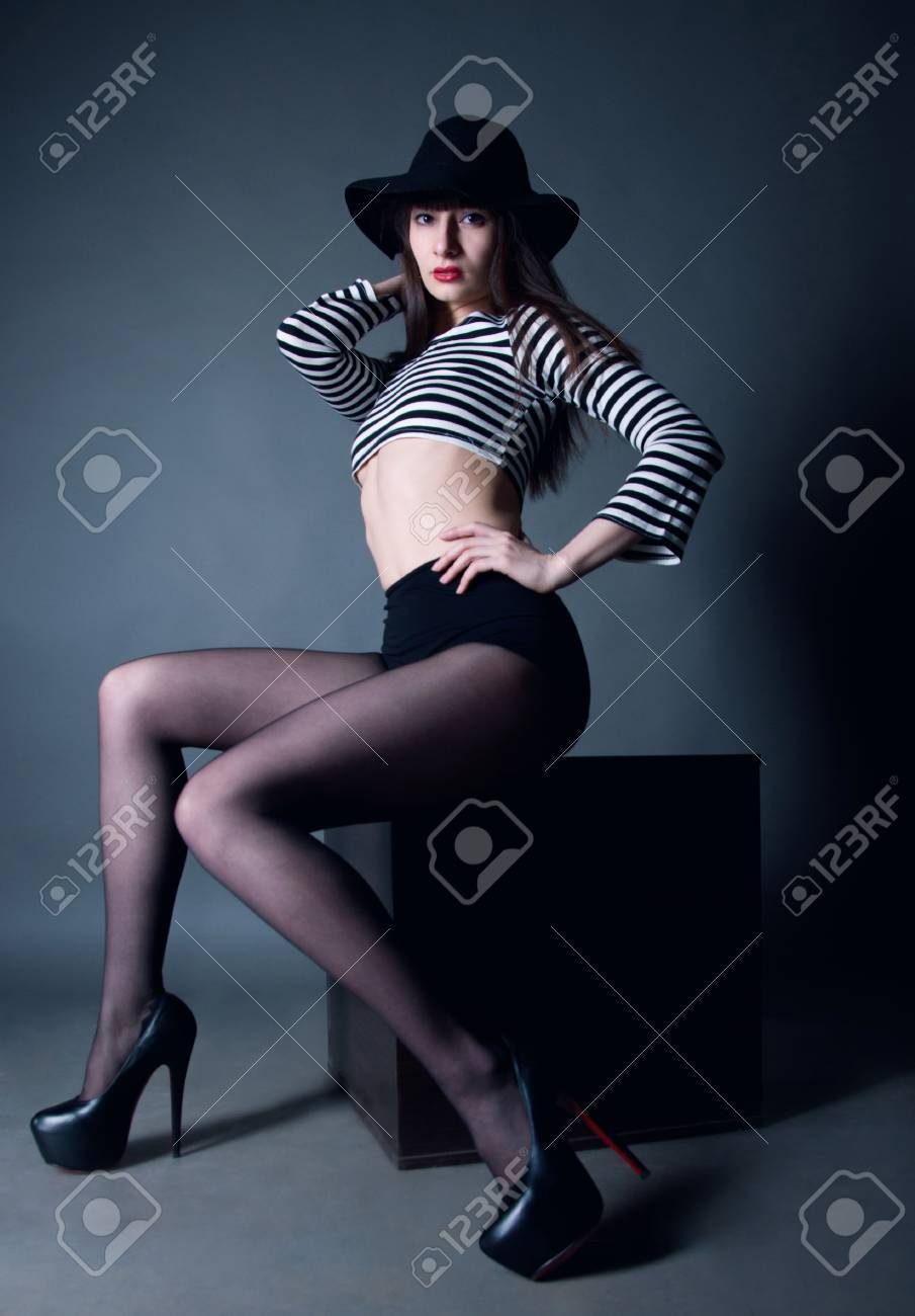 赤い唇と驚くほどスリムな長い脚 腰とカメラ目線の頭に腕を保持して キューブの上に座ってストッキング帽子 黒い短いストライプのトップスを着ているファッションでかなりブルネットの女性モデル 灰色の背景 の写真素材 画像素材 Image