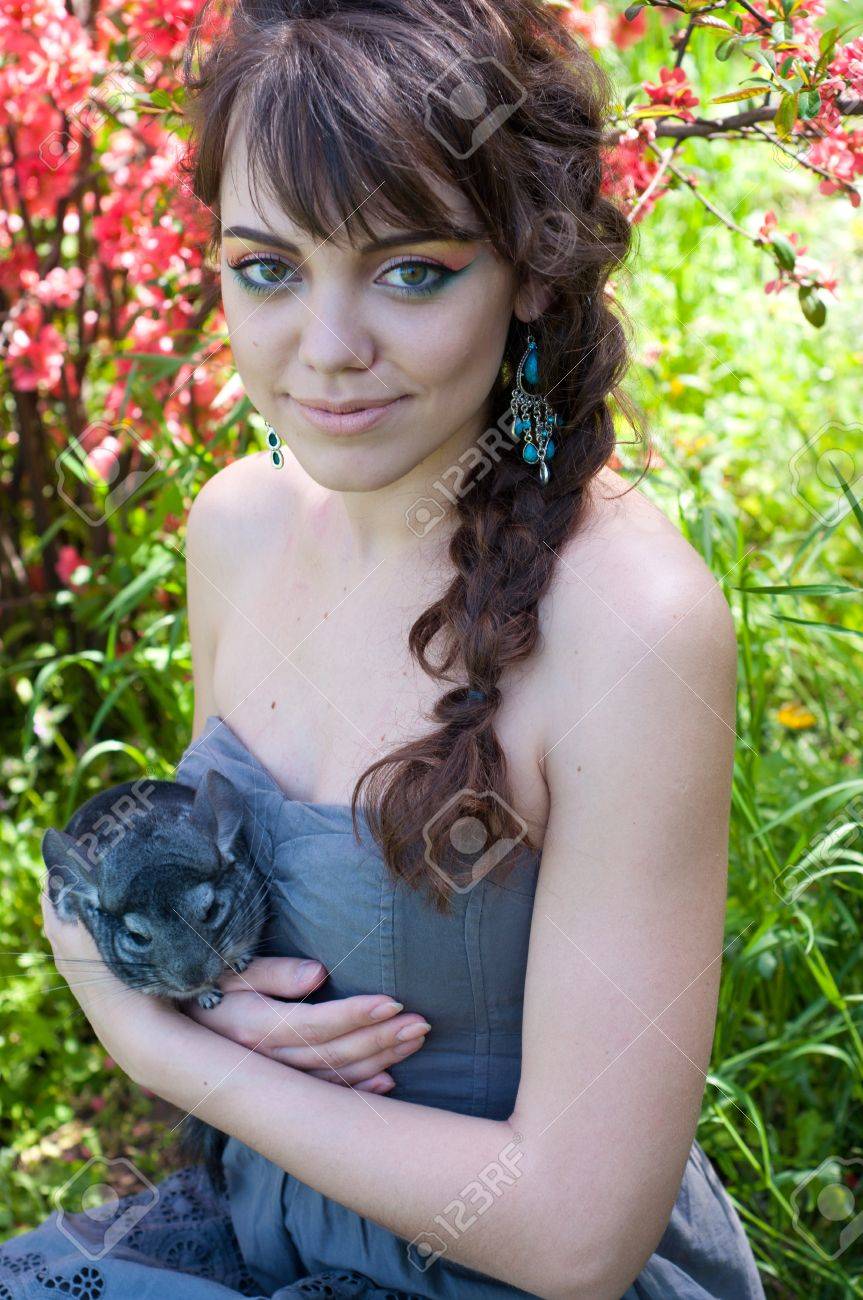 かなり若い女性モデルの夏のドレスを着て明るいカラフルなメイクアップと 青のイヤリング 彼女のペットのチンチラを手につかまって すぐそれを押すと 笑みを浮かべて カメラ目線します 開花木に対して の写真素材 画像素材 Image