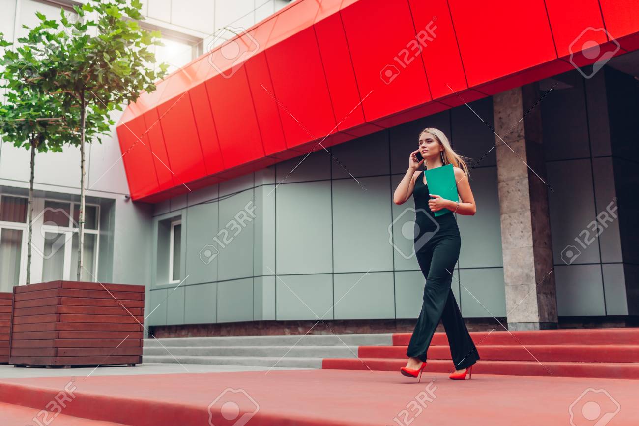 Young blonde businesswoman calling on smartphone and holding folder walking by business center in city. Business, communication, technology and people concept - 105584794 Young blonde businesswoman calling on smartphone and holding folder walking by business center in city. Business, communication, technology and people concept - 105584794