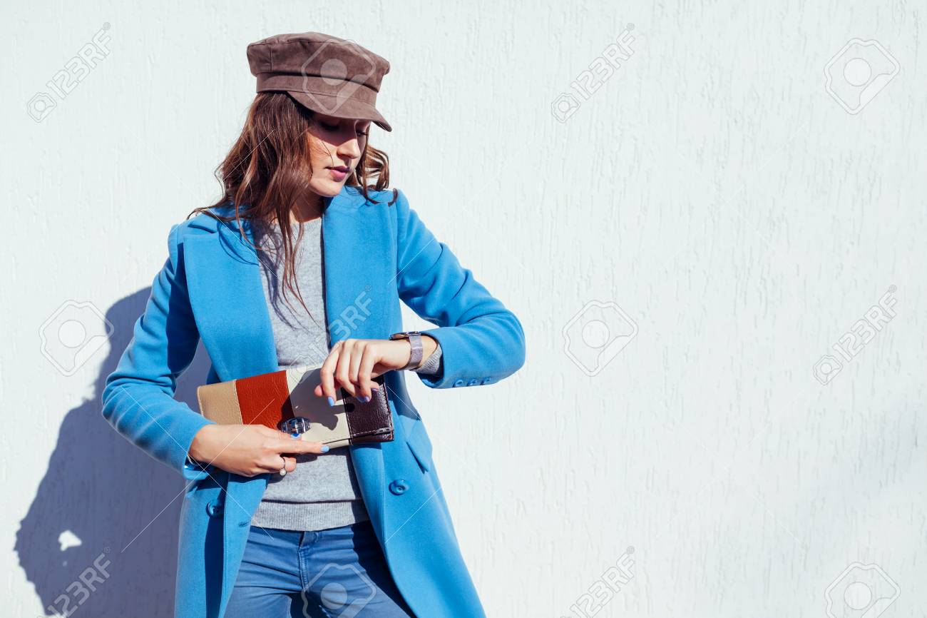 Young woman looking on watch, holding stylish handbag and wearing trendy blue coat and cap. Spring female clothes and accessories. Fashion - 119334044 Young woman looking on watch, holding stylish handbag and wearing trendy blue coat and cap. Spring female clothes and accessories. Fashion - 119334044